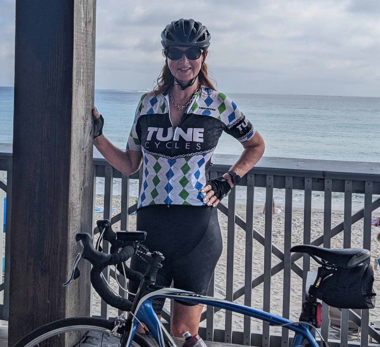 Tracy, 65, in cycling kit and helmet standing with her road bike on a boardwalk overlooking the beach in Delray Beach