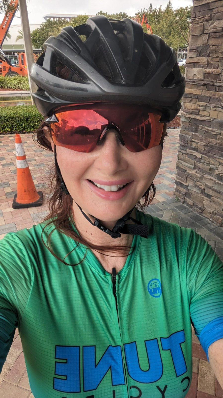 Tracy smiling after a ride in a green cycling jersey, helmet and orange-lens glasses