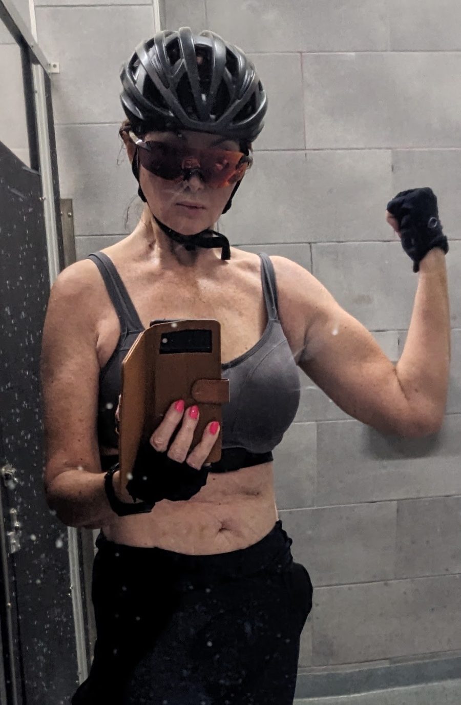 Tracy in a sports bra and cycling helmet flexing her arm in a locker room mirror